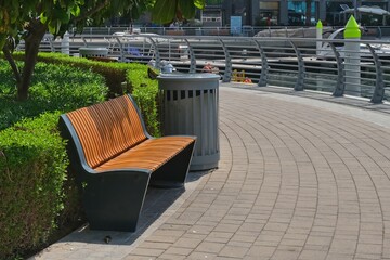 Empty contemporary wooden bench along trimmed green plant fence and curved pavement. Urban modern public vandal-proof furniture.Public city resting area design.