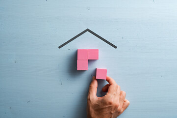Male hand assembling a house made of pink wooden blocks over blue background