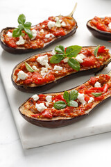 Baked eggplant stuffed with chopped tomatoes, sweet pepper, feta cheese and basil on marble cutting board and white background. Copy space.