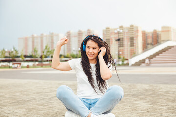 A smiling girl with dreadlocks in headphones is sitting on the floor in the park. A happy young woman relaxing with headphones, enjoying music. Space for copying.generation z