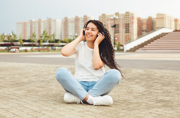 A smiling girl with dreadlocks in headphones is sitting on the floor in the park. A happy young woman relaxing with headphones, enjoying music. Space for copying.generation z