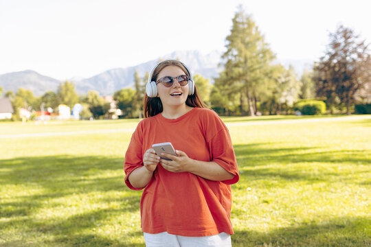Close up portrait of a lovely happy young woman in sunglasses listening to music through wireless earphones on nature background. Music lover enjoying music. Gigl choosing sound track on cellphone