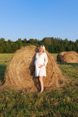 Naklejka na ściany i meble Young pregnant woman in a white dress stands barefoot at a haystack.