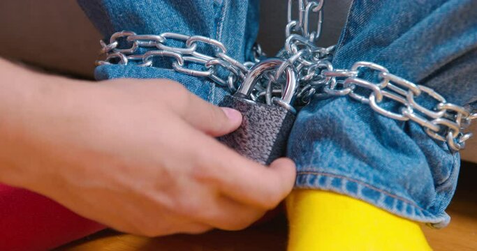 Men legs in jeans, bright socks with chain on ankles. Hand checks reliability of shackles, lock. Bright representatives of system who disagree with system, protests. Political prisoners, house arrest