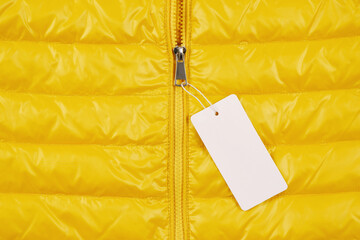 A bright background of a shortened yellow down jacket with an empty price tag on the zipper. A...
