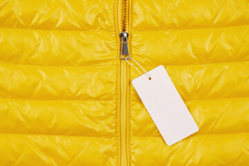 A bright background of a shortened yellow down jacket with an empty price tag on the zipper. A...