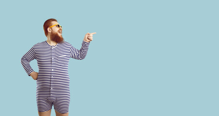 Funny guy in striped pajamas showing something on the right. Happy fat man wearing funny PJs or old retro swim suit standing isolated on blue background, pointing finger at copy space and laughing