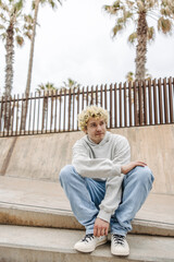 In full length young caucasian guy with blonde curly hair sitting at street. Calm male dressed casual outfits with palm trees in background. Concept summer vacation.