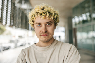 Close-up of calm young caucasian guy looking at camera with serious face on street. Curly blonde...
