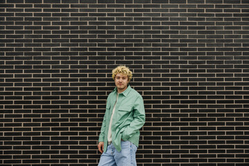 Fototapeta na wymiar Calm young caucasian guy dressed green shirt keeping his hand in pocket. Curly man standing near brick wall. Concept fashion, lifestyle.