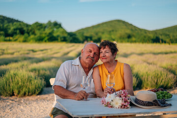an elderly couple in love, a man and a woman in the summer on a lavender plantation, sit at a table with glasses in their hands