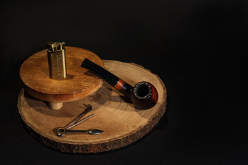 Tobacco pipe lying on a piece of wood with a pipe tamper and a lighter on a black background 