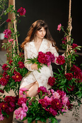 a beautiful young woman in a white dress sits on a swing in peony flowers. 