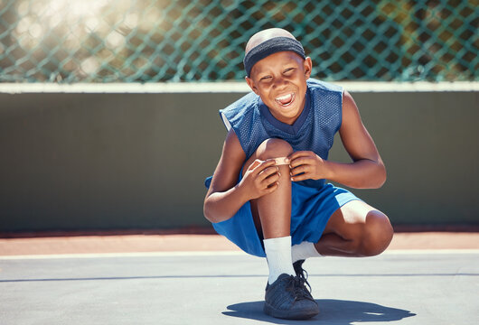 Child With A Band Aid On A Knee Injury From Sports Outside On A Basketball Court Touching His Bandage. Boy With A Medical Plaster Hurt By Accident While Training Or Practicing For A Game.