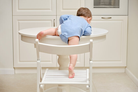 Toddler baby climbs on the kitchen white table. A child in danger climbs up on the furniture at the risk of falling. Kid aged one year and two months