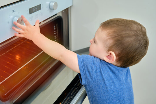 Toddler Baby Climbs Into A Hot Electric Oven. Child Boy Opens Oven Door In Home Kitchen. Kid Age One Year