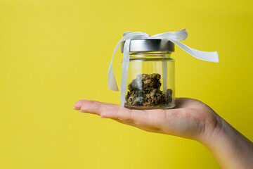 Female hand holding marijuana bud in glass jar with white ribbon on yellow background, close up. Concept of herbal and alternative medicine