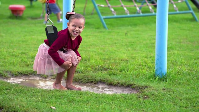 Ni&ntilde;a hermosa y feliz latina rubia colgada en un columpio divertida y disfrutando en el parque en un charco de agua y tierra y lodo al aire libre al exterior en un hermoso d&iacute;a