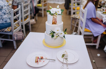 A large three-tiered white cake with a cut off piece on a plate with a fork and knife stands on a table in a restaurant. Food photography, wedding.