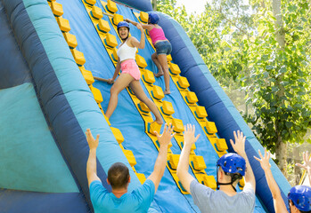 Happy laughing girl competing in climbing with wood poles on tall inflatable slide on adults bouncy...