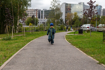 
a two-year-old toddler boy runs along the path in the park near the house on the street in autumn in a hat and overalls. Joyful and happy baby