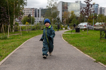 
a two-year-old toddler boy runs along the path in the park near the house on the street in autumn in a hat and overalls. Joyful and happy baby
