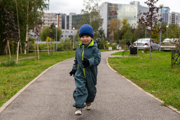 
a two-year-old toddler boy runs along the path in the park near the house on the street in autumn in a hat and overalls. Joyful and happy baby