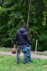 A little boy in a green jumpsuit hugs his dad's leg. Back view.