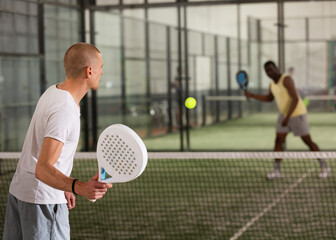 Rear view of man playing paddle tennis match on indoor court on blurred background of opponents. Sport and active lifestyle concept