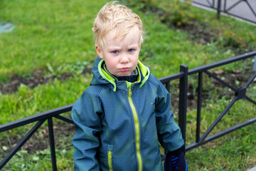 A two-year-old kid stands at the fence and pouted offendedly. Looks into the camera. Portrait. Autumn. No hat.