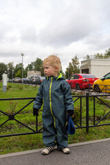 
A two-year-old kid stands at the fence in full growth and looks to the left into the distance, he took off his hat. Autumn.