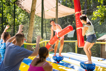 Fototapeta na wymiar Cheerful young girl with female friend having fun on inflatable gladiator fight arena in outdoor amusement park in summer..