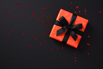 Red black Friday gift box wrapped black ribbon bow on dark background with confetti.