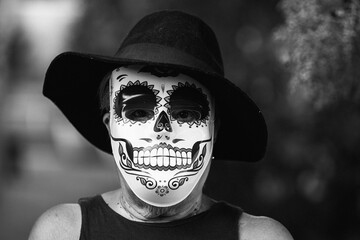 Black and white portrait of an elderly lady with a catrina mask and hat, celebrating Halloween and All Souls' Day, on the street. Celebration, costume, party and mask concept.