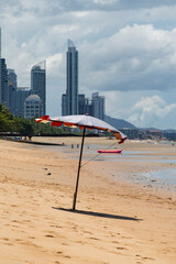 A parasol stands on the sand of a beach by the sea at low tide. City skyscrapers are in the background.