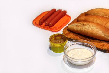 Defocus ingredients for making homemade hot dogs. Sausages in orange plate, fresh baked buns, mustard and sauce on white background.  Hotdog with different ingredients. Copy space. Out of focus