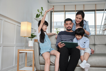 Happy Asian Thai family, parents, and children have fun using digital tablet together on sofa in home living room, a lovely leisure weekend, and domestic wellbeing lifestyle with internet technology.