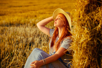 A beautiful young woman in a hat and a summer dress is sitting on a sheaf of hay in a field. Rural nature, wheat field, rest in the country, unity with nature