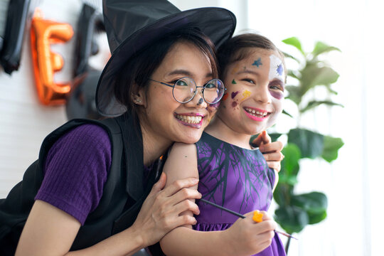 Mother and daughter wears witch Halloween costume and makeup their face together, hugging and smiling at camera at home,