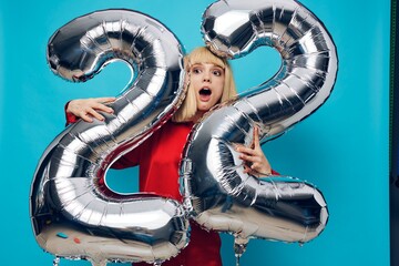 surprised, shocked woman in a red shirt stands with her mouth open on a blue background and holds inflatable balloons in the shape of the number twenty-two in silver color, hugging them with her hands