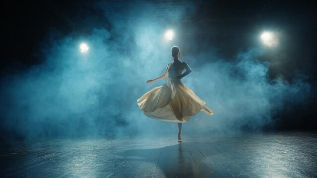 Graceful ballerina dances in light flowing dress on stage in pointlight in stage smoke. Ballet dancer performs dance elements of classical ballet