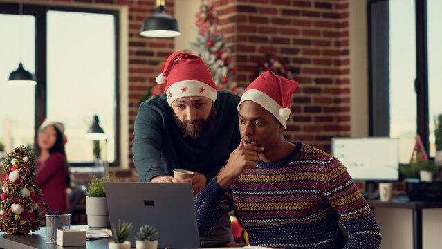 Multiethnic team of people analyzing report on computer during christmas eve festivity, wearing santa hats. Diverse men working together in office with xmas tree and seasonal ornaments.