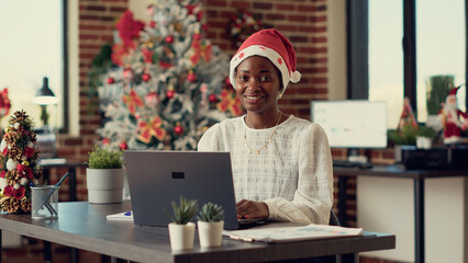Portrait of african american woman wearing santa hat at work, working with research data on laptop...
