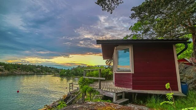 A red cottage by a lake in Sweden at sunset with a colorful sunset reflecting off the water - motion time lapse