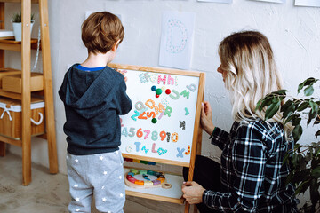 Kindergartener stand with back and learn english letters, alphabet and numbers on magnetic board. Educator sit near boy in playroom and check exercise. Reading and counting, study in daycare centre