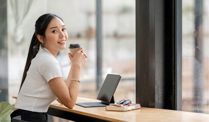 Smiling Asian businesswoman holding a coffee mug and laptop at the office. Looking at the camera.