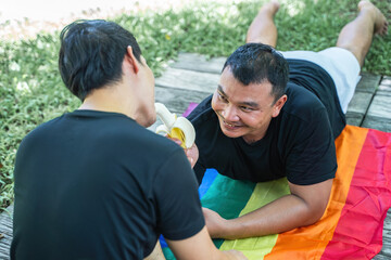 One of gay friends is holding a banana, giving it to taste his pal and laughing. Young gay couple having a romantic picnic in the park. lgbt, lgbtq, lgbtq+, homosexual