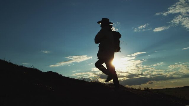 Dark silhouette of man in hat going uphill enjoying hiking in rural area at bright back sunset light. Determinated traveler walks on hill against illuminated blue sky with white clouds backside view