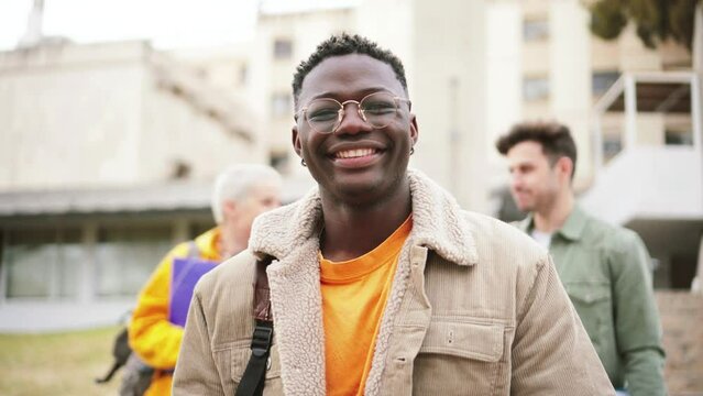 Young african american student with googles smiling and looking at camera standing at the high school, at the back ground a group of teenagers walking and laughing. Education concept. High quality 4k