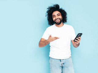 Handsome smiling hipster  model.Sexy unshaven Arabian man dressed in white summer t-shirt and jeans clothes. Fashion male with long curly hairstyle posing near blue wall in studio. Holds smartphone